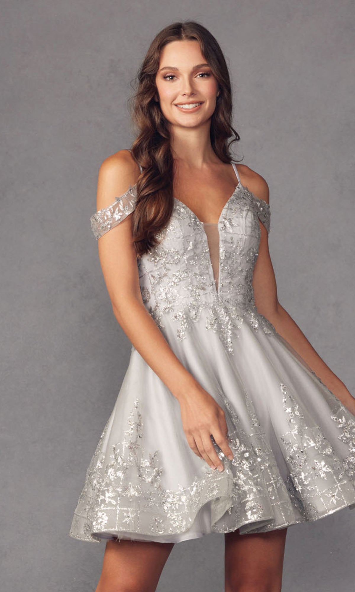A woman smiles while wearing the Glitter-Print Short Babydoll Homecoming Dress 903, featuring silver glitter fabric with floral sequin details, standing against a plain gray background.